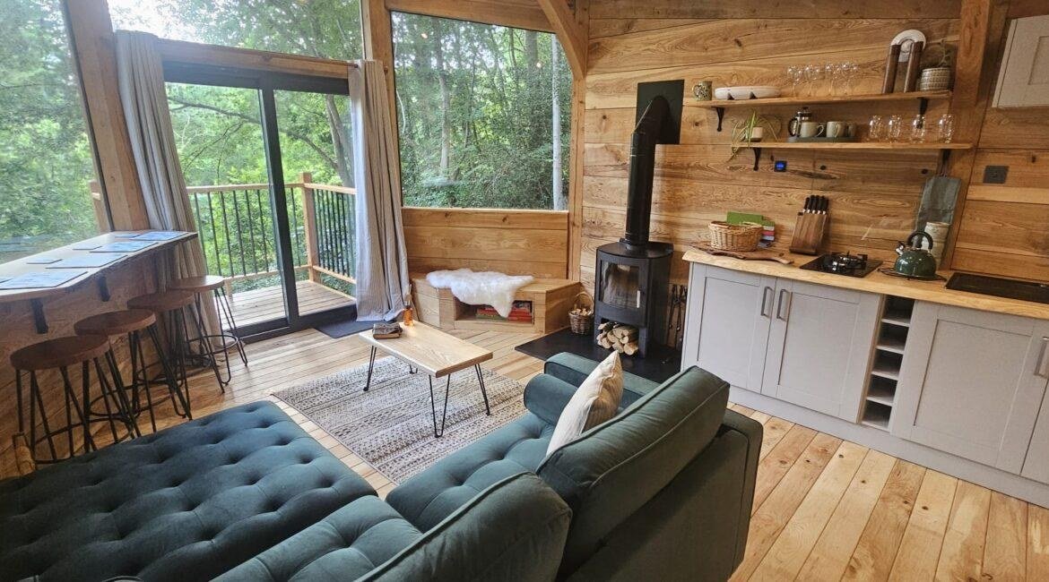 Open-plan interior of the Toad Hideout with wood burner, kitchen, sofa and large woodland windows at Tree Tops and Train Tracks in North Wales