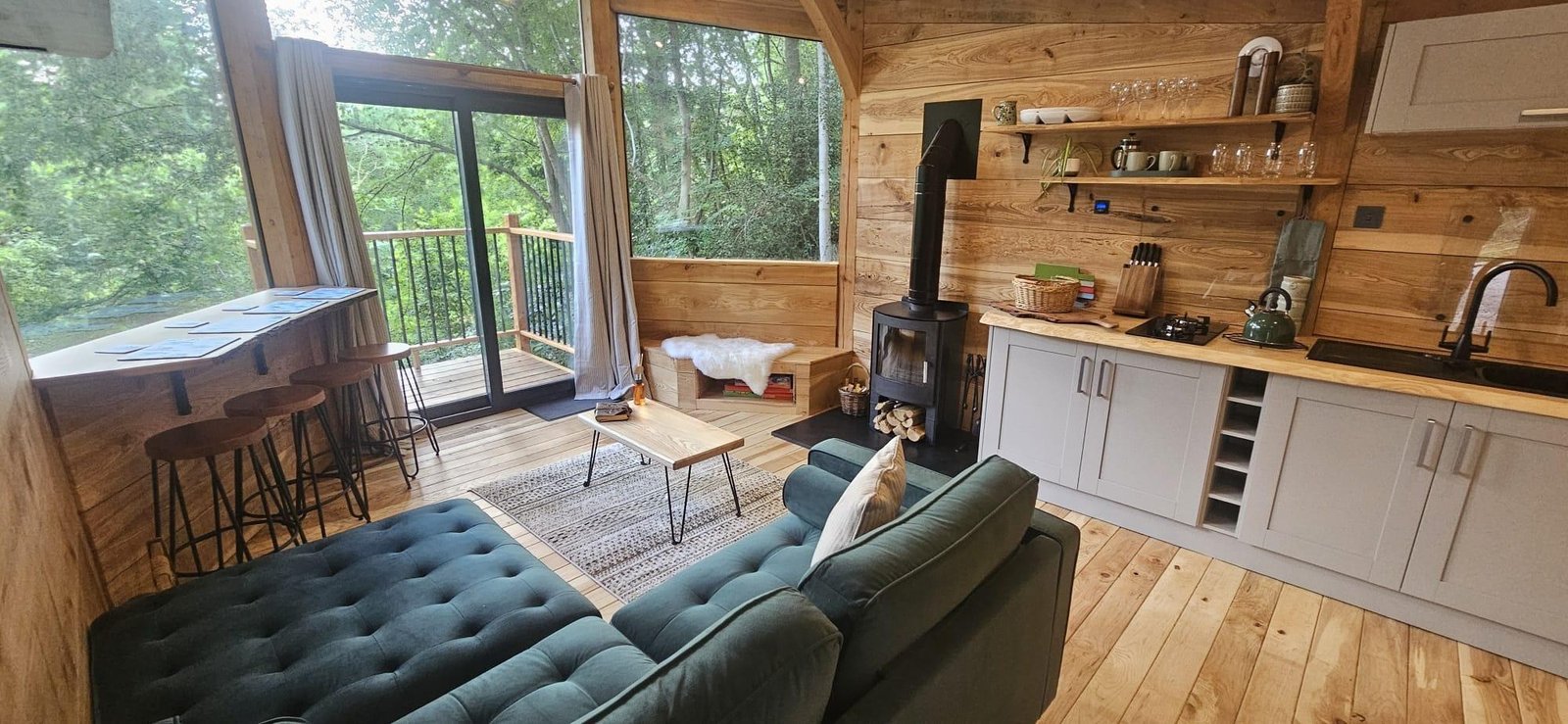 Open-plan interior of the Toad Hideout with wood burner, kitchen, sofa and large woodland windows at Tree Tops and Train Tracks in North Wales